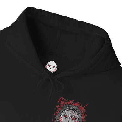 Folded Hooded Sweatshirt of Billy the Puppet staring forward with a sinister grin, his black suit and red bow tie framed by chains, gears, and rusted metal. A dim industrial setting fades behind him as ominous red light highlights his face. The composition emphasizes psychological tension, horror, and the iconic image of the puppet who announces Jigsaw’s deadly games.